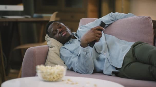 Keskipitkä laukaus nuori laiska afrikkalainen amerikkalainen mies makaa sohvalla television edessä yöllä, vaihtaa kanavaa sitten nukahtaa kaukosäädin käsissä - Materiaali, video