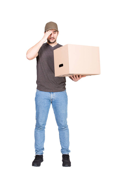 Confused delivery man, full length portrait, holding a cardboard box while keeps a hand to forehead, isolated on white. Puzzled courier has problems delivering package, messed up the parcels - Photo, Image