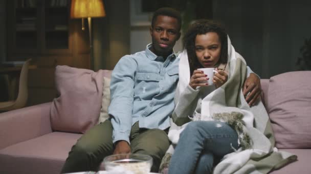 Lassú-mozgás zoom POV fiatal szerető afro pár néz filmet együtt ül a kanapén éjszaka. Nő rosszul érzi magát ivás forró tea borított kockás, míg a férfi megöleli őt - Felvétel, videó