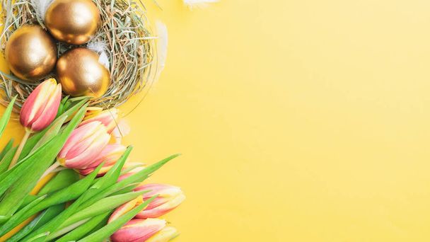 Easter egg. Happy Easter decoration: Golden eggs in basket with spring tulips, white feathers on pastel yellow background. Traditional decoration in sun light. Top view - Photo, Image