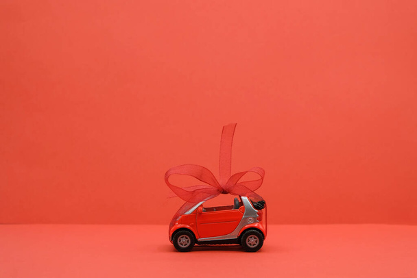 Černobílý obraz. Miniaturní hračka červené auto s červeným lukem na červeném pozadí. Dárek, překvapení, nebo současný koncept. Sny se stávají skutečností. Kopírovat prostor. - Fotografie, Obrázek