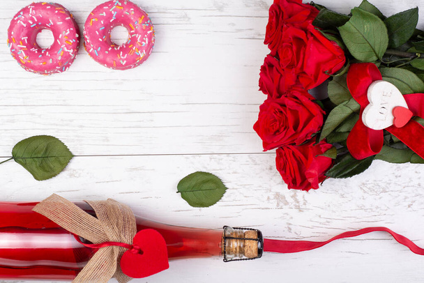 バレンタインデーの背景。フレームの赤いバラ、ボトルシャンパン、装飾的な心、ピンクのドーナツ、素朴なリボンは、木製の表面に。おめでとうございます。女性の日の贈り物。お祝いセット.. - 写真・画像