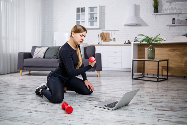 Junge sportliche Frau in Fitnessbekleidung in einem modernen Zuhause, die Online-Unterricht von einer Fitness-Website am Laptop nutzt und zu Hause trainiert. Sport zu Hause während der Quarantäne - Foto, Bild