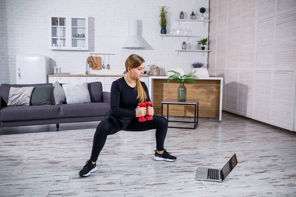 Junge athletische Frau in Turnkleidung, schwarzem Top und Leggings in einer Wohnung mit Online-Workout von einer Fitness-Website auf einem Laptop und Sport zu Hause. Fitness zu Hause während der Quarantäne - Foto, Bild
