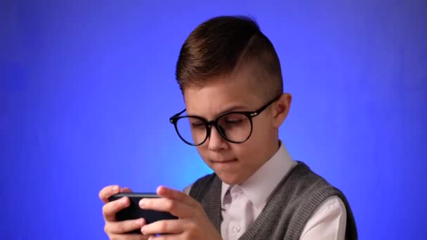 Jóképű iskolás fiú szemüveggel játszik egy játékot okos telefonon a kék háttér. - Felvétel, videó