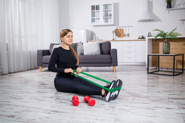 Uśmiechnięta młoda kobieta ćwicząca z elastyczną opaską podczas treningu fitness w domu. Koncepcja zdrowego stylu życia. Sport w domu podczas kwarantanny - Zdjęcie, obraz