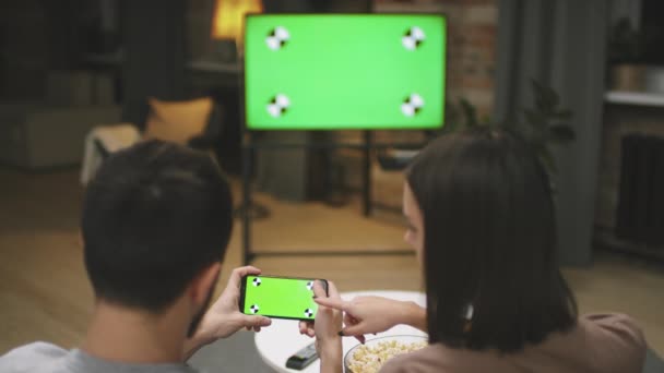 Back-view slow-motion medium close-up van jong stel zitten op de bank in de woonkamer naar voetbalwedstrijd kijken en samen spelen op smartphone - Video