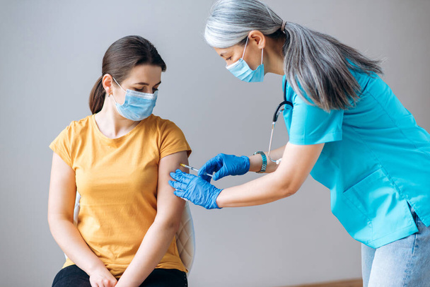 Médico de pelo gris femenino en guantes protectores y máscara médica, inyecte la vacuna inyectable para estimular la inmunidad de la paciente femenina para la infección por coronavirus. Concepto de vacunación contra el Coronavirus - Foto, imagen