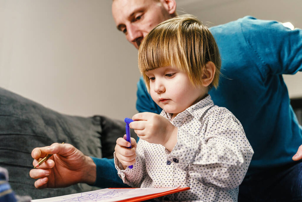 Small caucasian boy sitting on the sofa bed with his father playing with felt pen - Son and father spending time together learning and drawing at home - family time parenthood and childhood concept - Photo, Image