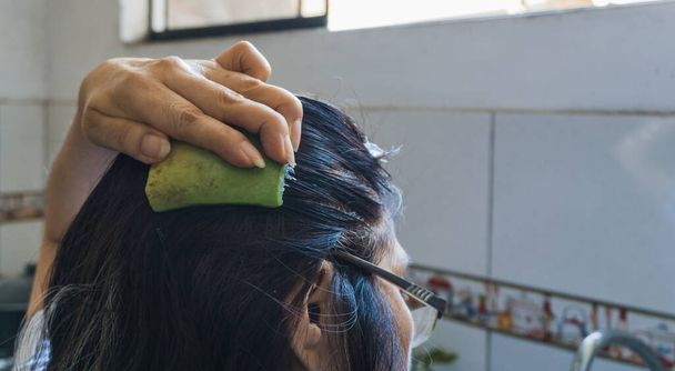 αγνώριστη ενήλικη γυναίκα που απλώνει φλοιό αλόης στα μαλλιά της σε μια κουζίνα με μη εστιασμένη βρύση και φόντο πλακιδίων - Φωτογραφία, εικόνα