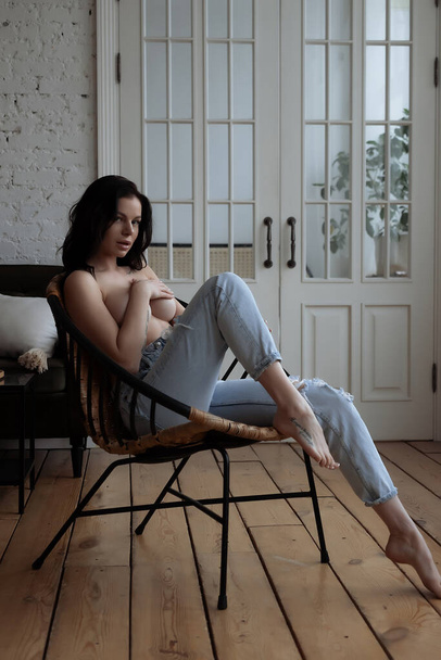Чуттєва молода жінка в джинсах покриває голу груди рукою, розслабляючись один в кріслі в затишній кімнаті вдома
 - Фото, зображення