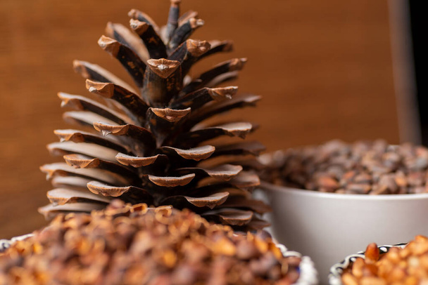A large open pine cone next to a ceramic bowl. They contain pine nuts, shells and peeled nuts.  - Photo, Image