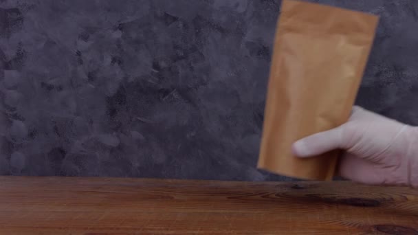 Ruční ochranné rukavice drží hnědé kraft papírové sáček s kávovými zrny dřevěné pozadí 4K mockup. Balení pro dodávání potravin šablony zboží. Balení s uzávěrem okno čaj listy váha produktu. - Záběry, video