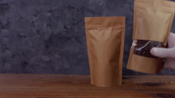 Ręka w gumowych rękawiczkach posiada brązowe torby papierowe torby makieta z ziaren kawy pokazuje kciuk w górę jak drewniane tło Opakowanie żywności dostawy towarów szablon Kraft opakowanie z zapięciem herbata liście waga produktu - Materiał filmowy, wideo