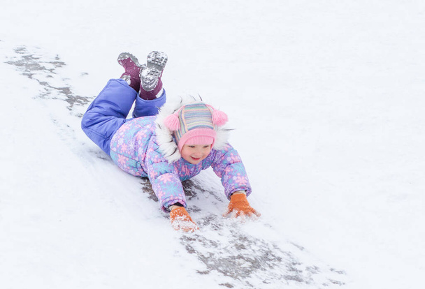 A little pretty girl in a purple overalls slides down an ice slide, winter children's games, a child in the park in winter. - Photo, Image