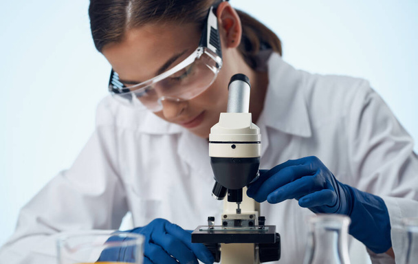 Laborassistentin mit Mikroskop auf dem Tisch und Brille auf der Gesichtsflüssigkeit in einem Kolben - Foto, Bild