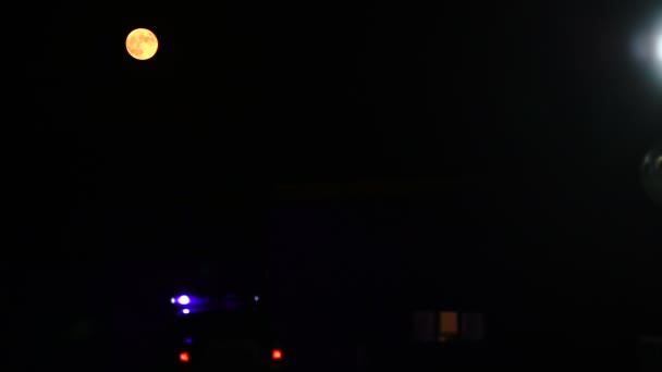 Gecenin karanlığında büyük turuncu ay, binanın yanında duran polis arabasının çok renkli ışıkları, sokak lambasının altında yürüyen bir adamın silueti - 4K - Video, Çekim