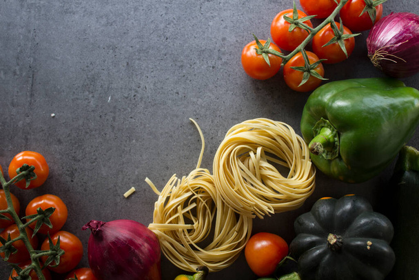Fresh pasta ingredients on a table. Top view photo of raw tagliatelle pasta, cherry tomatoes, red onion, zucchini, garlic, pepper and salt. Dark grey background with copy space. Healthy eating concept - Photo, Image