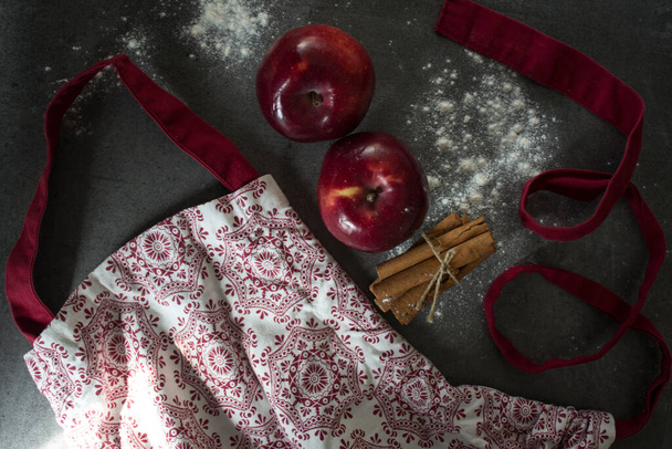Фото с кухонного стола. красный фартук, яблоки, коричные палочки, мука на темно-сером текстурированном фоне. Делаю торт. Концепция здорового питания.  - Фото, изображение