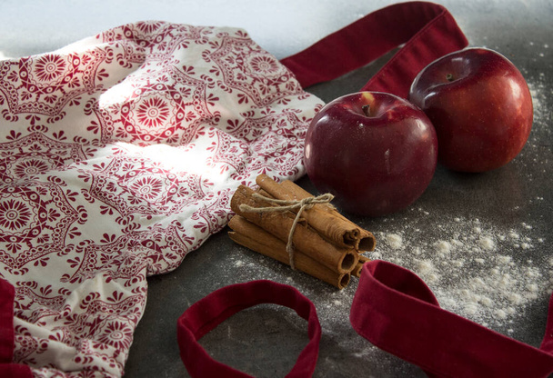 Zamknij zdjęcie czerwono-białego fartucha, dwa piękne jabłka i paluszki cynamonowe na stole kuchennym. Widok z góry zdjęcie składników szarlotki.  - Zdjęcie, obraz