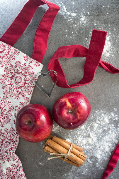 Ποδιά κουζίνας, κόκκινα μήλα, αλεύρι, ξυλάκια κανέλας πάνω όψη φωτογραφίας. Φτιάχνω μηλόπιτα. Σκηνή οικογενειακής ζωής. - Φωτογραφία, εικόνα