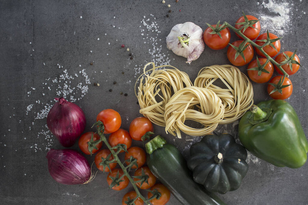 Fresh pasta ingredients on a table. Top view photo of raw tagliatelle pasta, cherry tomatoes, red onion, zucchini, garlic, pepper and salt. Dark grey background with copy space. Healthy eating concept - Photo, Image
