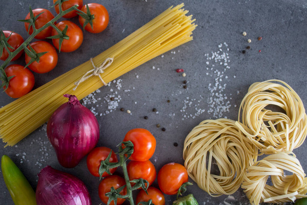 Verse pasta ingrediënten op een tafel. Bovenaanzicht foto van rauwe tagliatelle pasta, kerstomaten, rode ui, courgette, knoflook, peper en zout. Donkergrijze achtergrond met kopieerruimte. Gezond eten concept - Foto, afbeelding