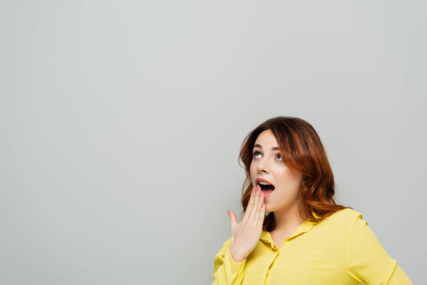 mujer emocionada cubriendo la boca abierta mientras mira hacia otro lado aislado en gris - Foto, Imagen