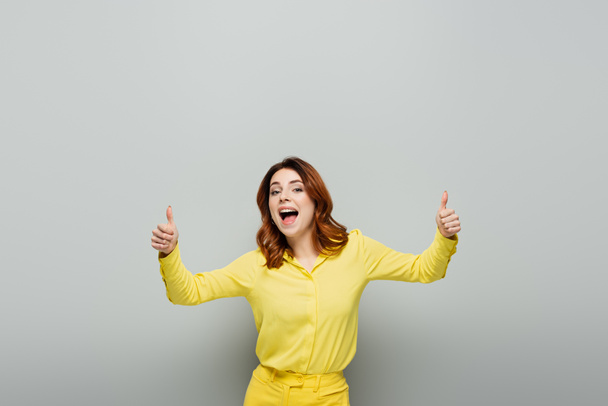 joyful woman showing thumbs up while standing with open mouth on grey - Photo, Image