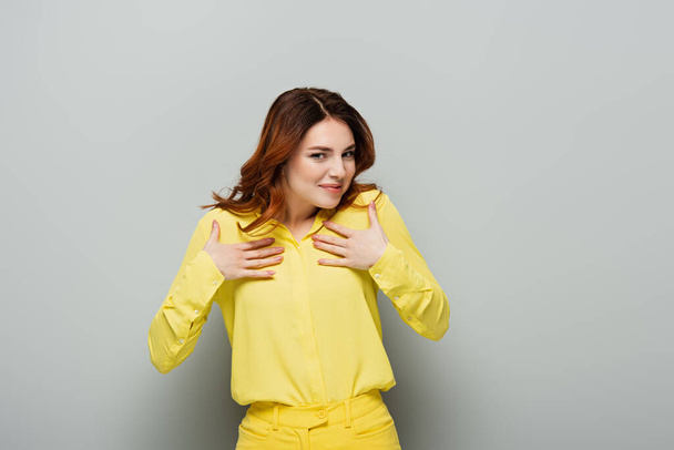 pleased woman in yellow shirt holding hands on chest while looking at camera on grey - Foto, imagen