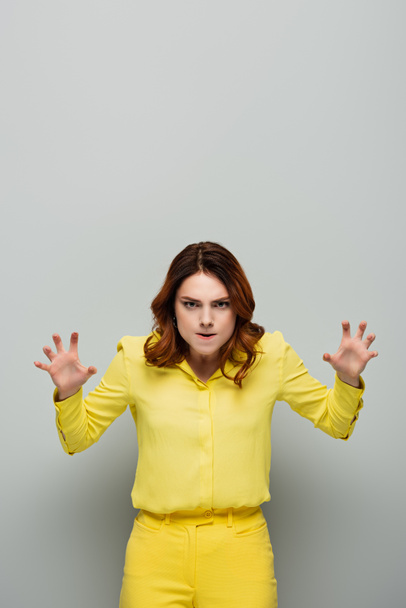 Wütende Frau im gelben Hemd blickt in die Kamera und zeigt angsteinflößende Geste auf grau - Foto, Bild