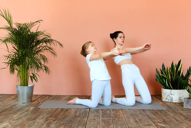 Vista lateral de la mujer sonriente y la niña practicando yoga mientras está de pie en la estera en la acogedora sala de estar y mirando hacia otro lado - Foto, Imagen