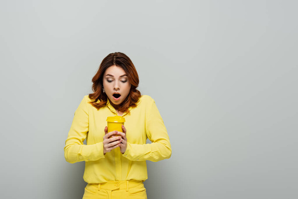 shocked woman in yellow blouse holding hot coffee in yellow paper cup on grey - Foto, afbeelding