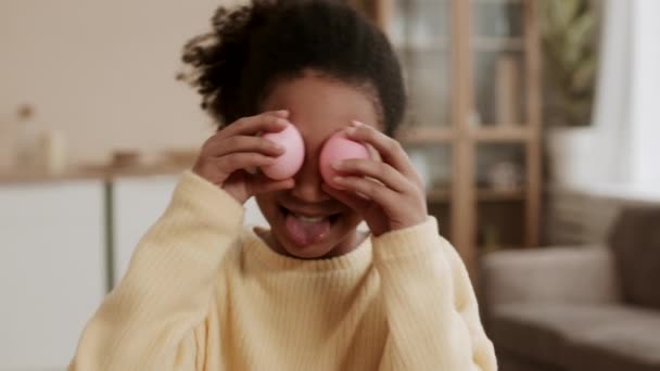 Medio primer plano de la alegre chica africana haciendo cara divertida, mostrando la lengua, sosteniendo huevos de color rosa en los ojos, luego quitando las manos y mirando a la cámara - Metraje, vídeo