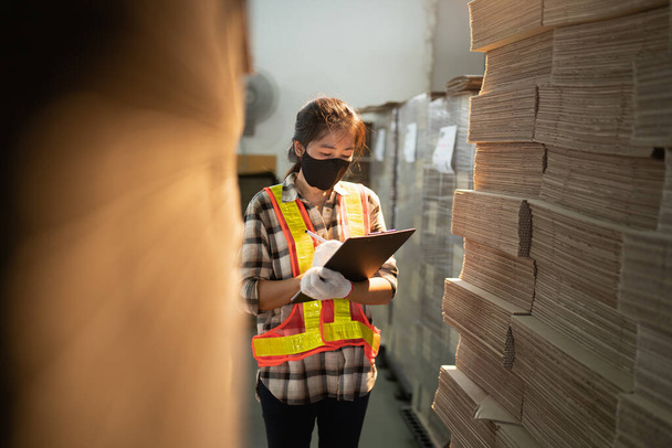Travailleuse d'entrepôt travaillant dans un entrepôt avec presse-papiers, Asie fille portant un masque facial tout en faisant une vérification de stock dans l'atmosphère de travail de l'usine. - Photo, image