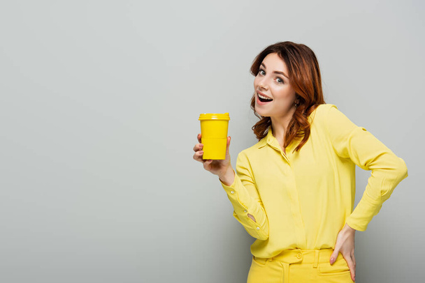 cheerful woman holding yellow paper cup while standing with hand on hip on grey - Foto, afbeelding