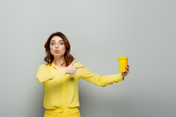 excitée femme regardant caméra tout en pointant vers tasse en papier jaune sur gris - Photo, image