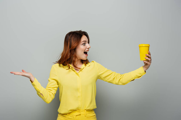 curly excited woman pointing with hand while holding coffee to go on grey - Photo, image