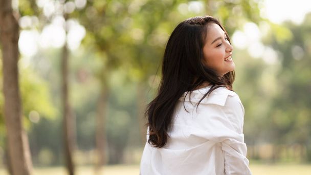 白いシャツを着た若いアジア人女性の姿をカジュアルに笑顔で描き、公園の自然光の中で人生を楽しむ、リラックスした瞬間、自然を楽しむ、気をつけ、新鮮なものを感じる. - 写真・画像