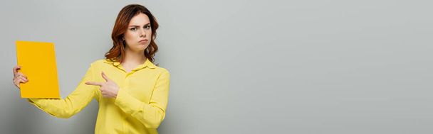 serious woman in yellow blouse pointing at notepad on grey, banner - Photo, Image