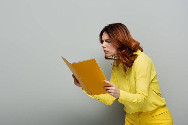 шокована жінка в жовтій блузці дивиться в блокнот на сірому
 - Фото, зображення
