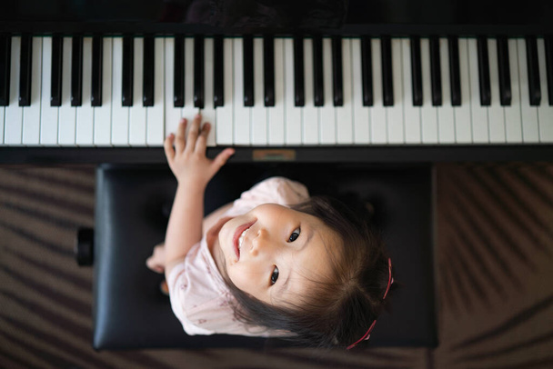 Asian toddler girl look up with smiling while playing piano, exploring music instrument, performance, early childhood music education, leisure activity, top view - Photo, Image