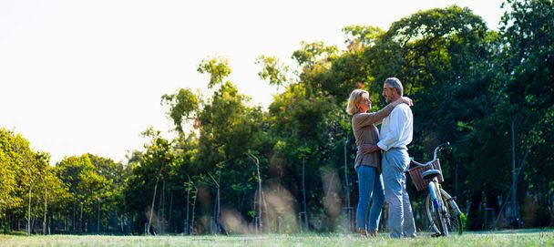 自然の秋の日差しの庭で自転車の横に立っている白人の高齢者のカップルのパノラマとお互いを見て大切で愛を感じ、高齢者の愛と健康の概念退職生活. - 写真・画像