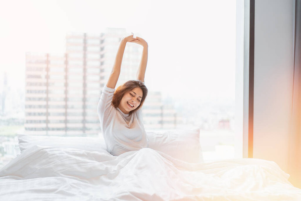 Ébresztő jelenet fiatal nő fényes reggel friss és pihentető nap. kényelmes életmód a hálószobában, kilátással a városra a reggeli napsütésben. - Fotó, kép