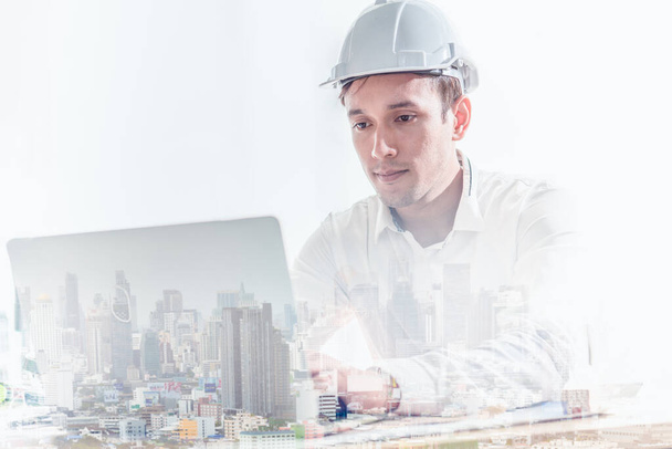Подвійне експонування інженера-будівельника в твердій шапці з ноутбуком і видом на місто
. - Фото, зображення