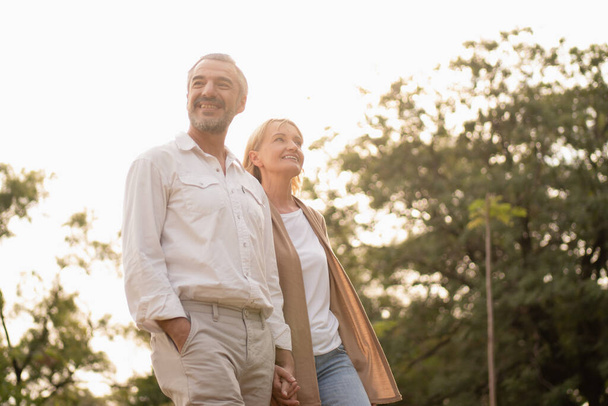 Senioři aktivní kavkazský pár držící se za ruce vypadají šťastně a bezstarostně v parku v odpoledním podzimním slunci s kopírovacím prostorem, šťastně v důchodu manžel, starší zdravý životní styl, šťastné výročí. - Fotografie, Obrázek