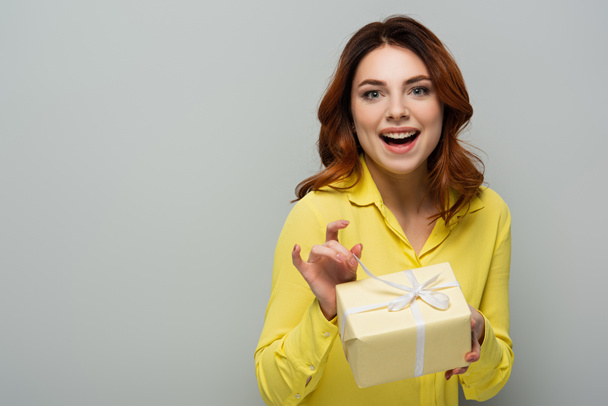 cheerful woman looking at camera while opening gift box on grey - 写真・画像