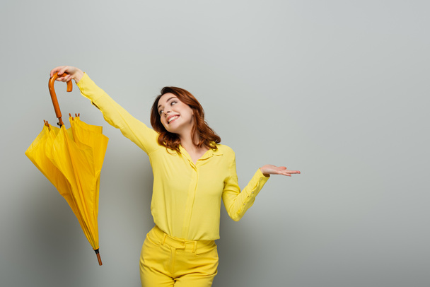 cheerful woman in yellow shirt standing with open arm while holding yellow umbrella on grey - Photo, Image