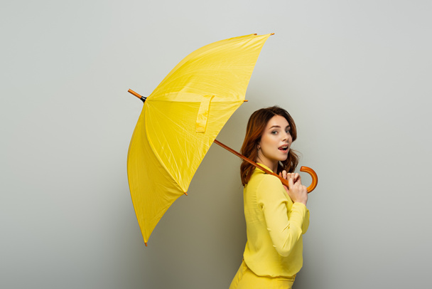 coquettish woman sticking out tongue while posing with yellow umbrella on grey - Photo, Image