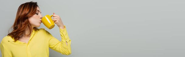 kręcona kobieta w żółtej bluzce pijąca herbatę na szarości, baner - Zdjęcie, obraz